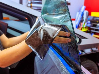 Worker technician applies window tint film to vehicle in workshop setting, focusing on precision quality.